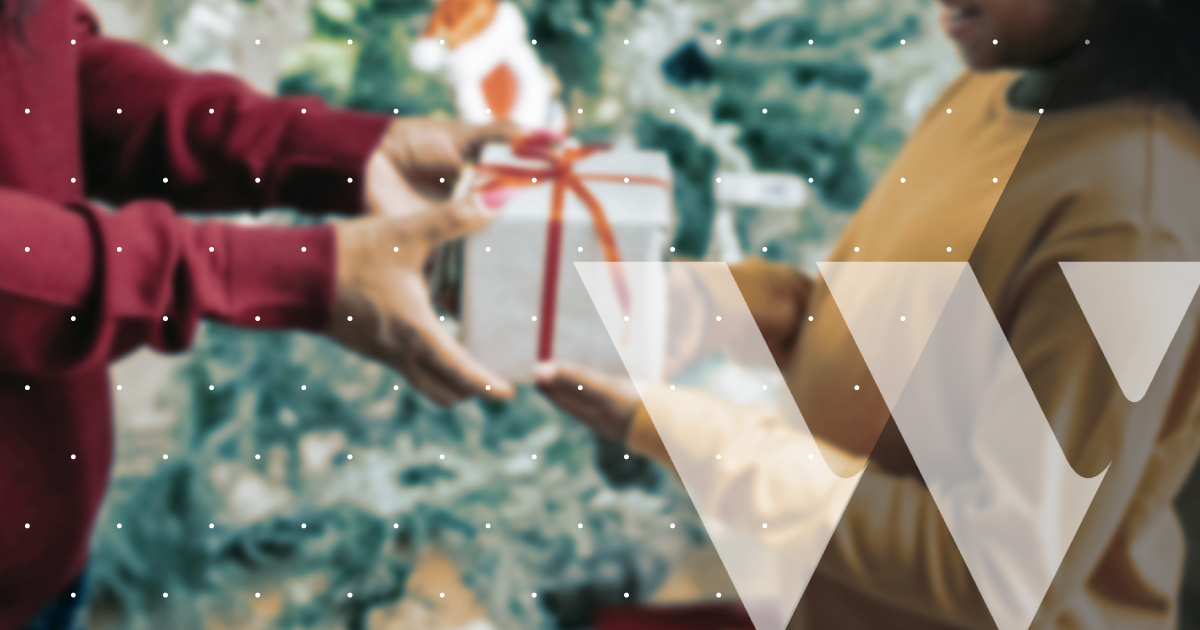 Rectangular image of one person handing a holiday gift to another person. On the bottom right of the frame, there is a white W, and there are tiny white polka dots throughout the image.