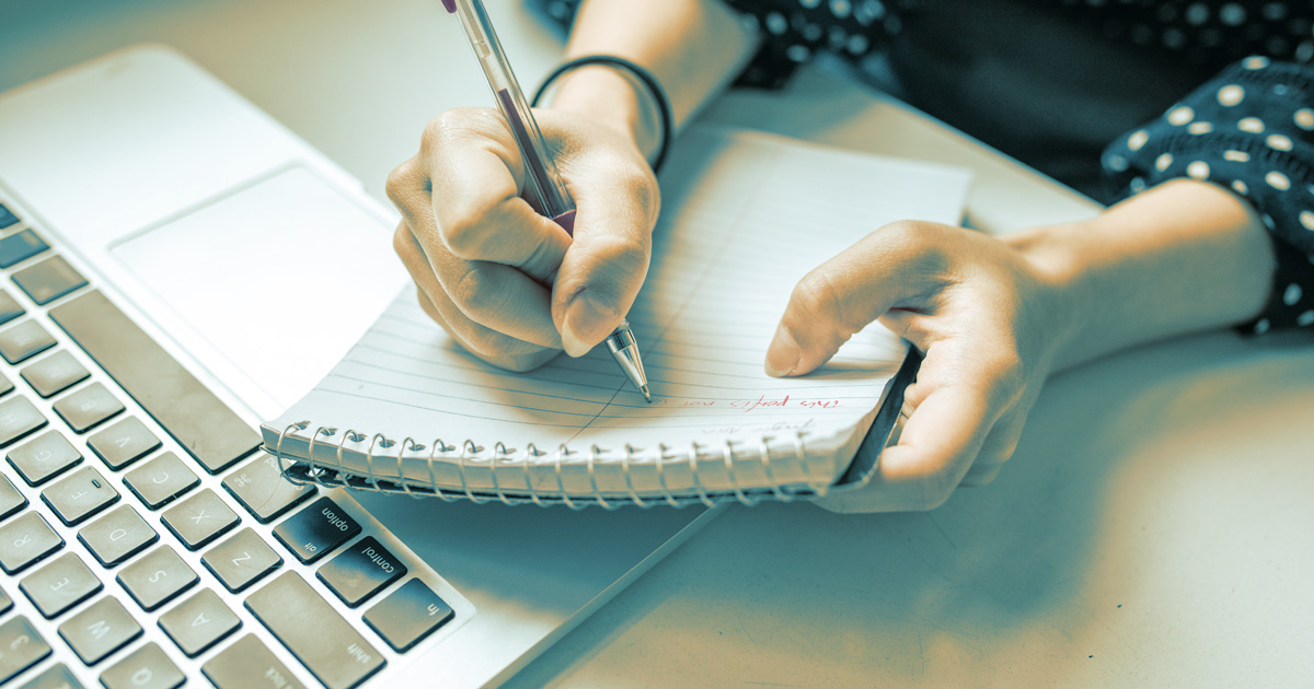Rectangular image of someone writing on a notepad. A keyboard is also visible.