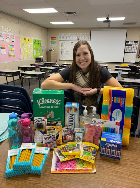Rectangular image. Teacher in a classroom smiling in front of a pile of various school supplies.